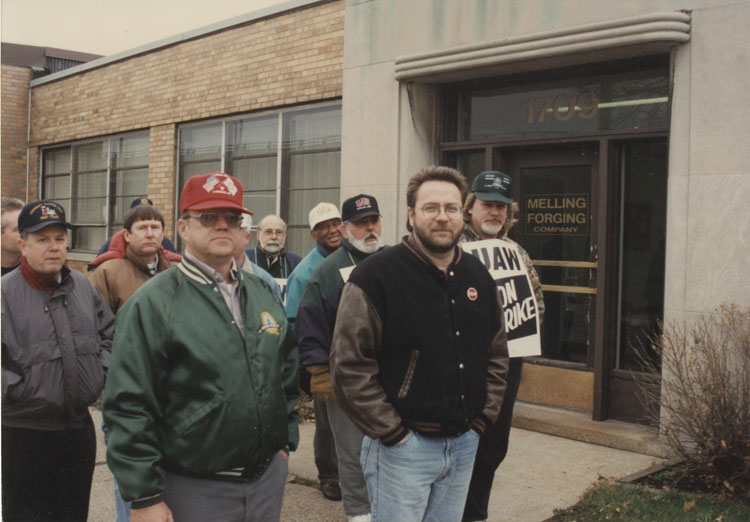 image of Melling Drop Forge in Lansing, MI., site of a strike against Melling by UAW local 724 approximately 2001-2002.  The subjects in the photo are members of UAW 724, UAW 602 and some Flint area UAW members supporting of the strikers.  Subjects are, from left to right:  Doug Taylor (mostly hidden), Jim Keller, Ken Michaud, hidden behind Michaud – Unknown, Gerald Taylor, hidden behind Taylor – Unknown, Unknown, Reuben Burkes (UAW Region 1-C Director, Unknown, Garry Bernath, Unknown.